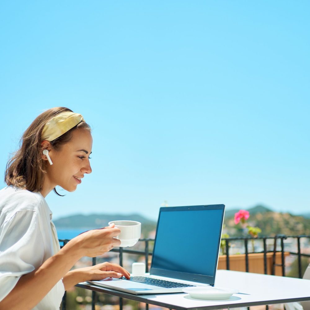 Adult beautiful woman using laptop computer typing emails sending messages drinking coffee enjoying working from Mediterranean resort cafe